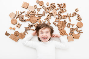 Boy, child laid on his back playfully smiling. Laid surrounded by wooden handmade toys. Wooden Letters, Shapes and Planets.