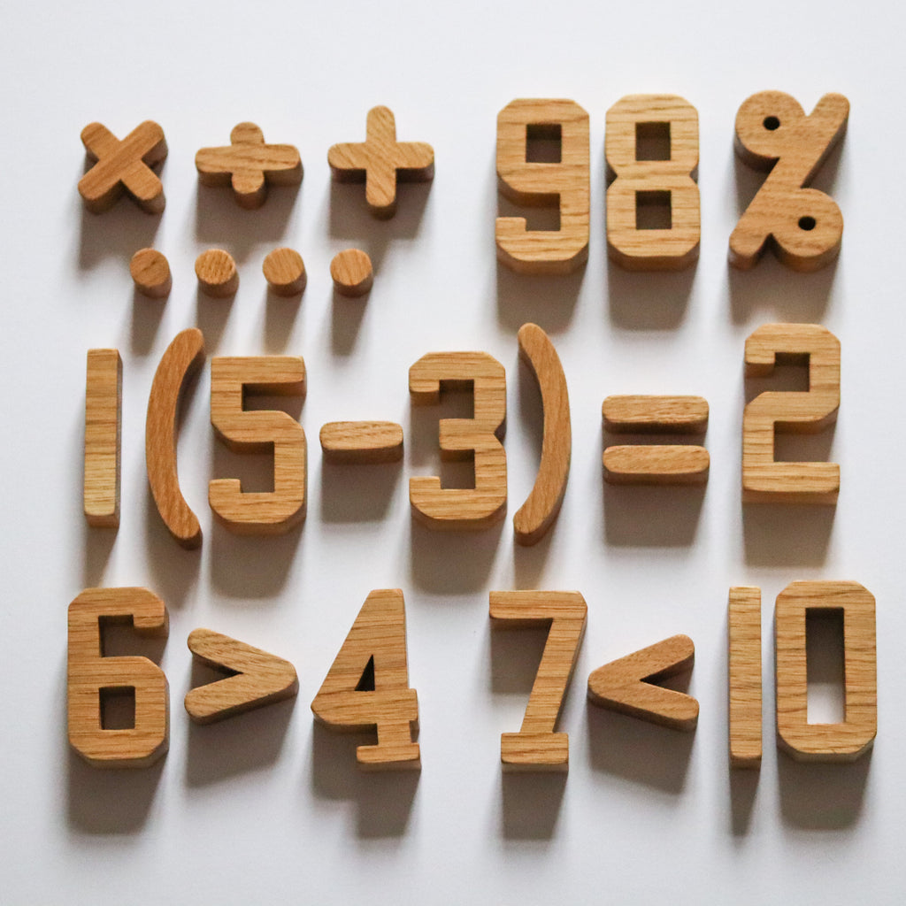 Wooden oak numbers and symbols toys. Arranged in equations. 6>4 7<10 on the bottom row. 1(5-3)=2 on the middle row. 98% and the symbols ×÷+ .... on the top row