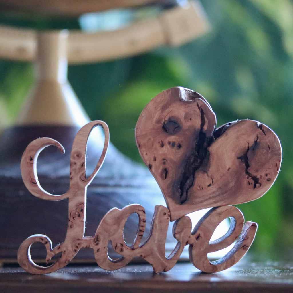 Wooden small script decor item spelling the word love. A burr elm heart balanced on the v and e with a wooden stand and globe and plant in the bacground 