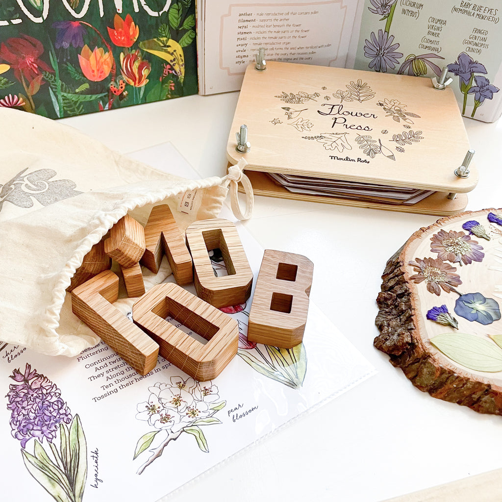 Wooden oak letters pouring out of a cotton bag spelling the word bloom. Flower press in the background and pressed flowers on a log slice.