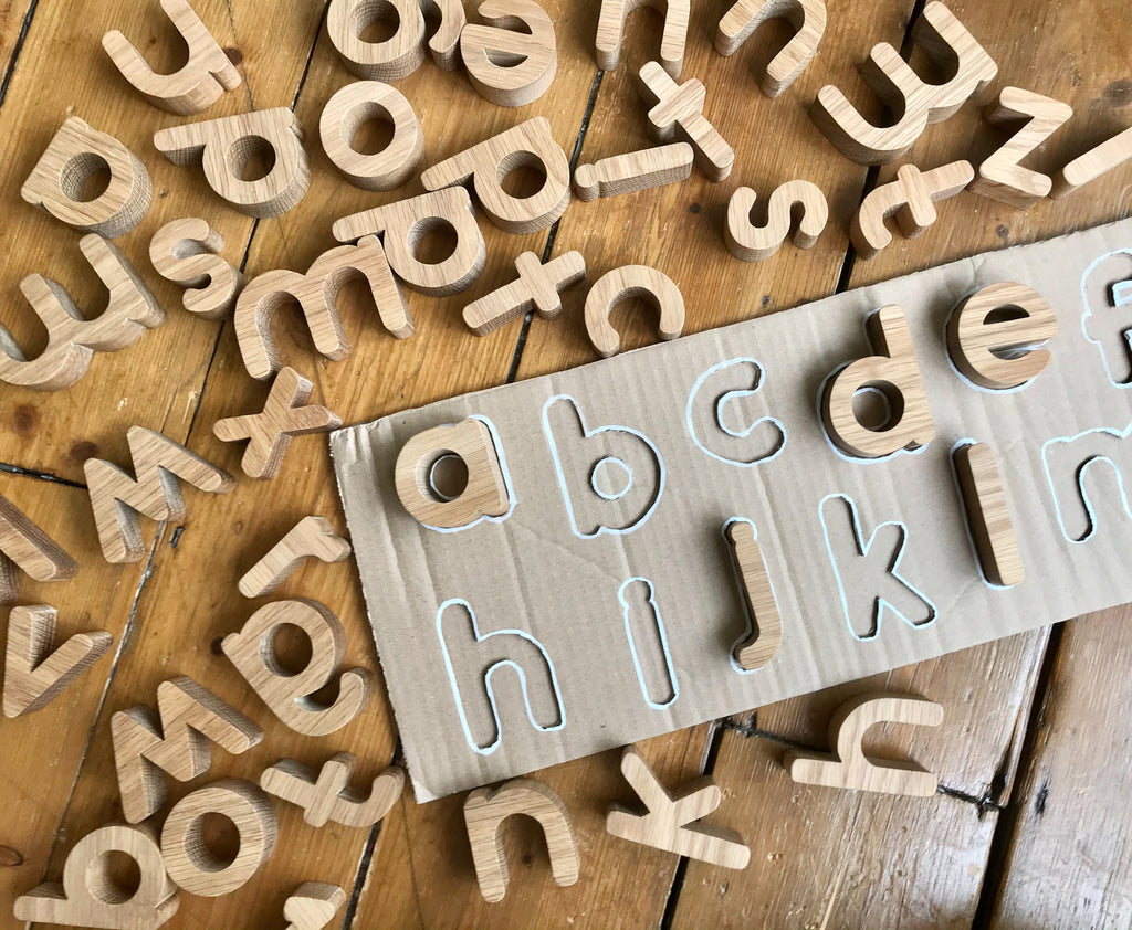 Handmade Wooden Oak Lowercase Alphabet Toy, scattered across wooden floor. A cardboard puzzle with some letters fit into place.