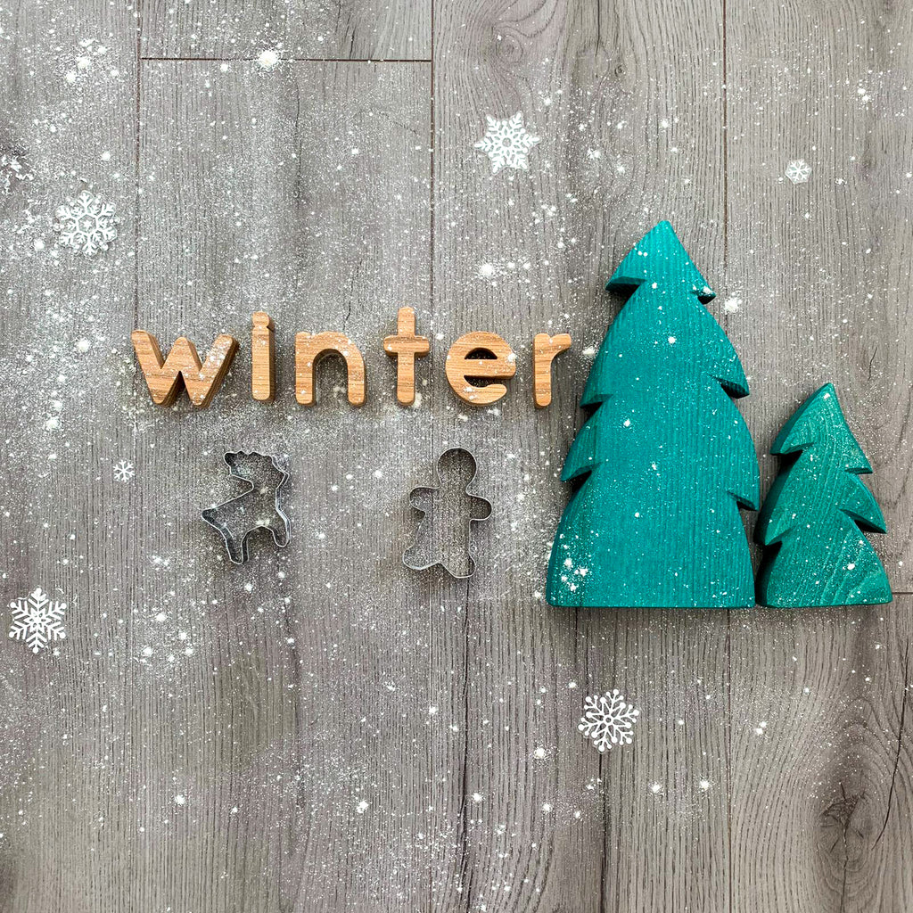 handmade wooden oak alphabet letters toy used to spell the word winter on a wooden background with a dusting of white powder and images of snowflakes. Next to a wooden green coloured tree toy.